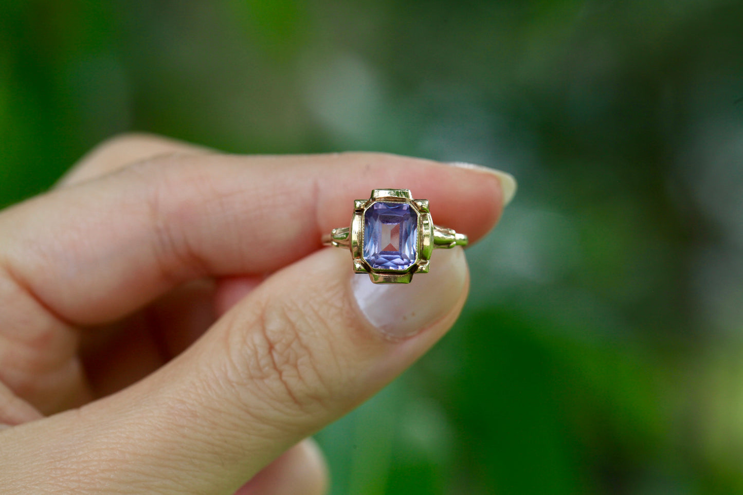 Vintage 10K yellow gold ring with an emerald-cut lavender amethyst, held between fingers against a blurred green background.