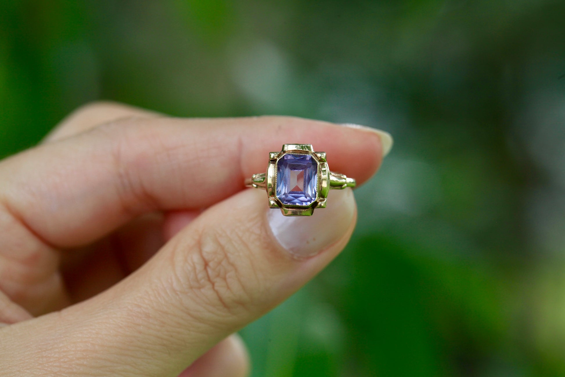 Vintage 10K yellow gold ring with an emerald-cut lavender amethyst, held between fingers against a blurred green background.