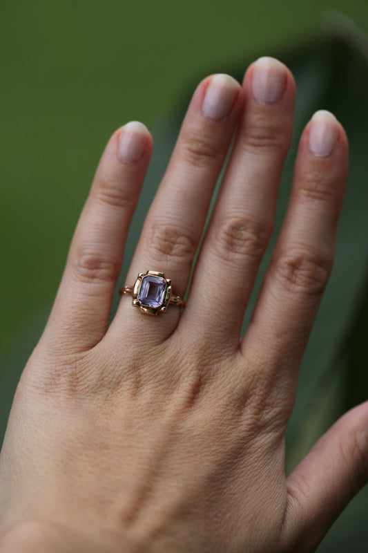 Vintage 1950s 10K yellow gold ring with an emerald-cut lavender amethyst, worn on a hand against a blurred green background.