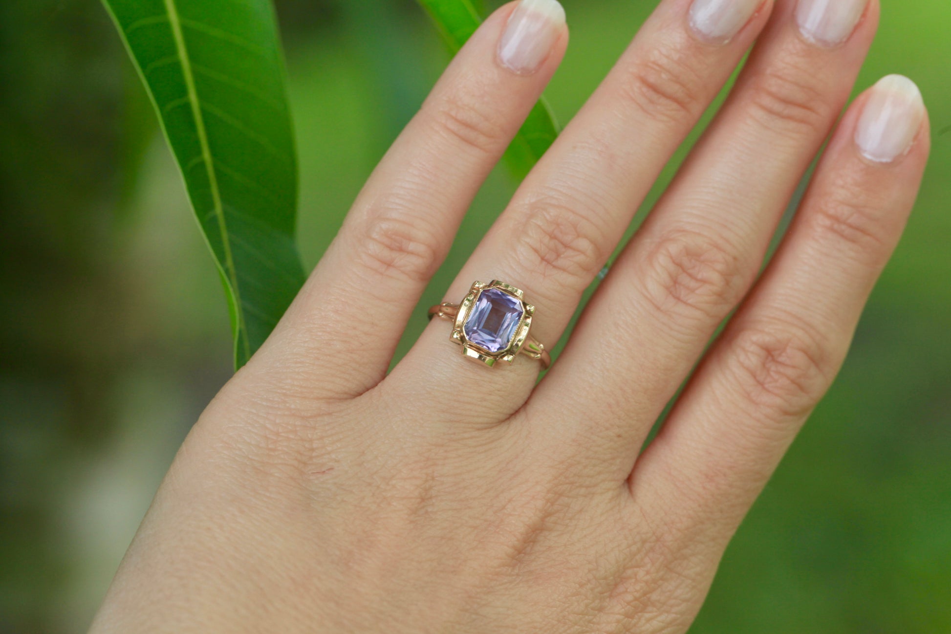 Vintage mid-century 10K yellow gold ring with an emerald-cut lavender amethyst, worn on a hand against a green leafy background.