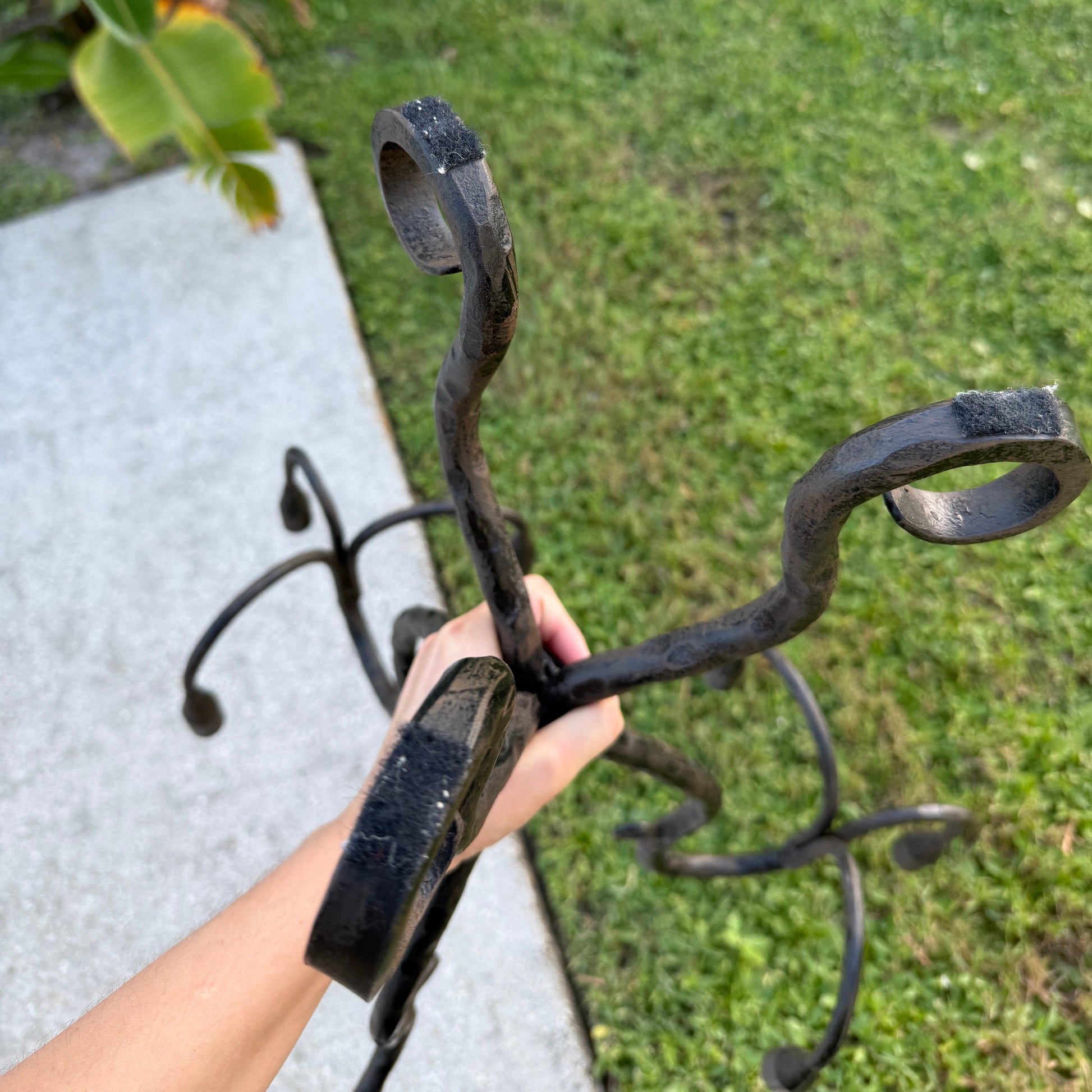 Hand holding a wrought iron candelabra outdoors, showing curved arms and textured black iron against a grassy background.