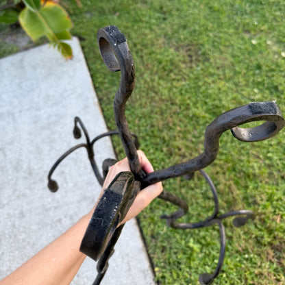 Hand holding a wrought iron candelabra outdoors, showing curved arms and textured black iron against a grassy background.