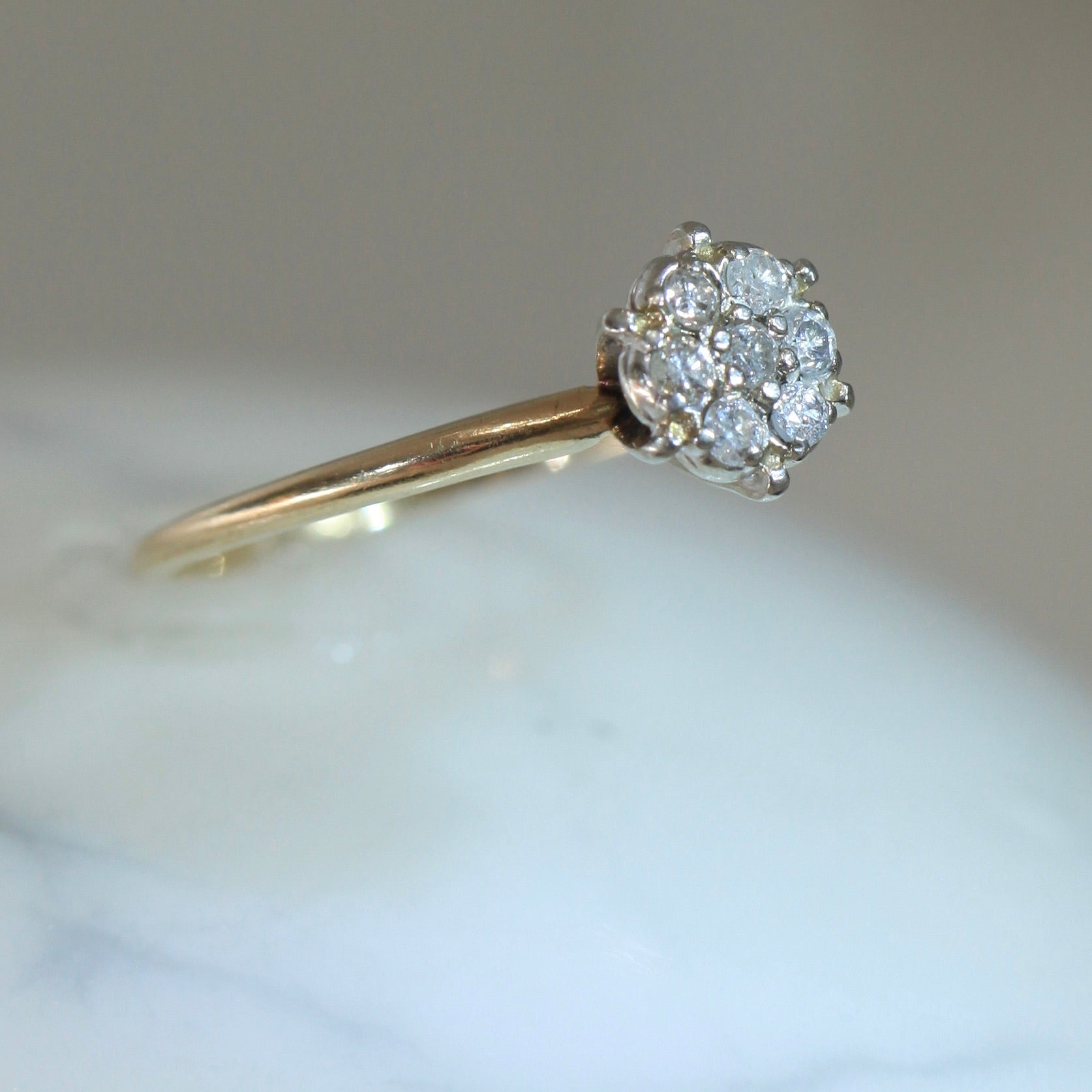 Angled view of 1950s vintage 10K two-tone gold engagement ring with seven-diamond floral cluster, slim yellow gold band on white marble surface