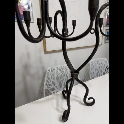 Wrought iron candelabra standing on a white tabletop, showing curved arms, candle cups, and sculptural tripod base.