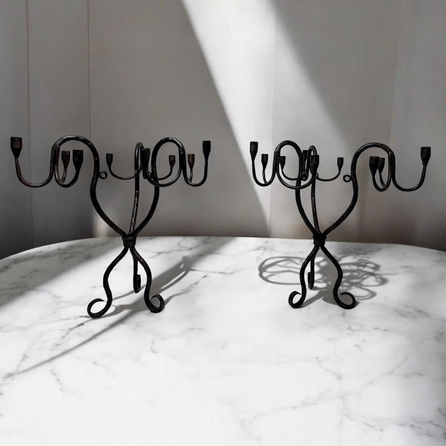 Pair of vintage hand-wrought iron candelabra with scrolling forged arms, displayed on a white marble surface in soft natural light.