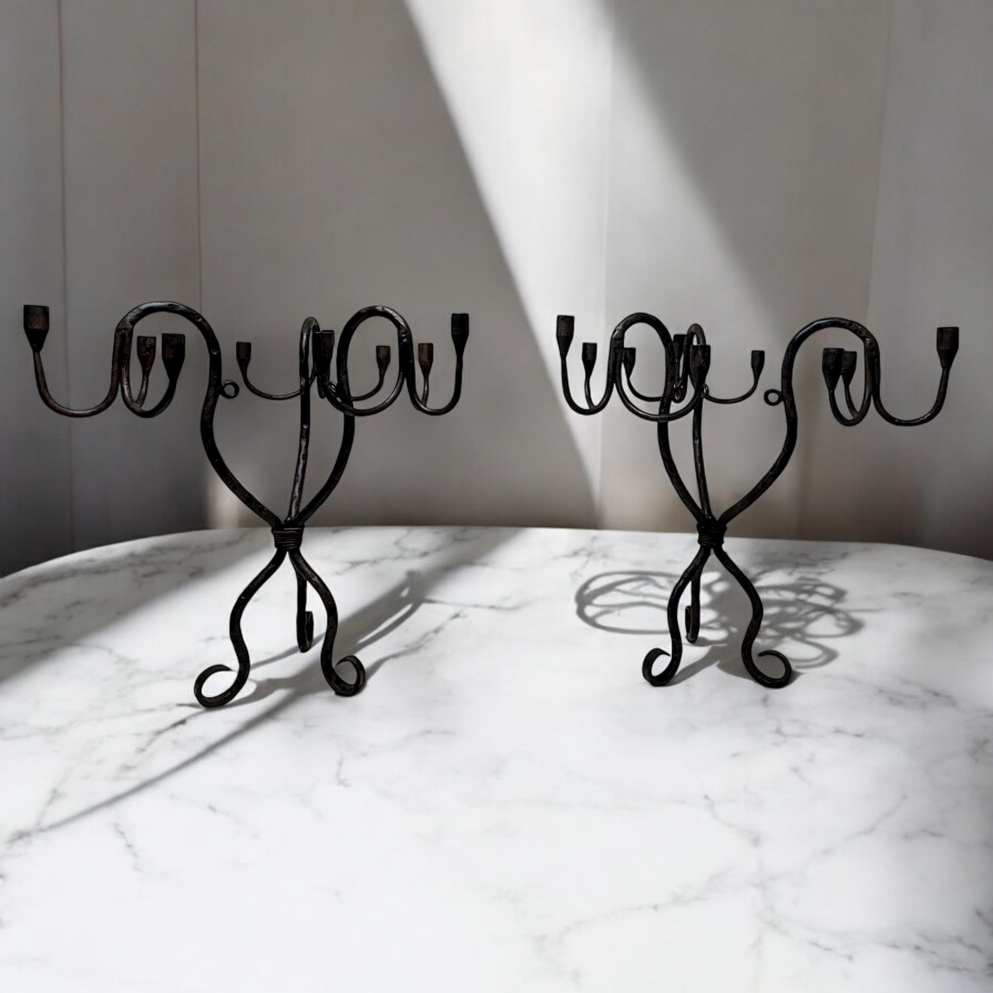 Pair of vintage hand-wrought iron candelabra with scrolling forged arms, displayed on a white marble surface in soft natural light.