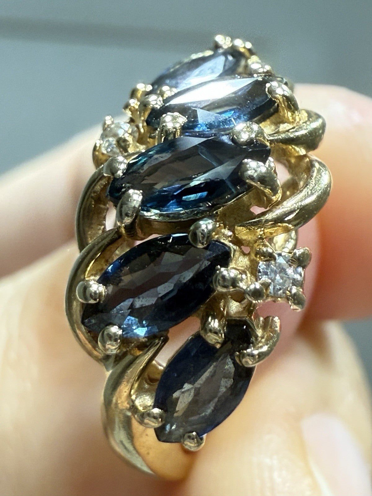 Close-up of vintage 1960s 14K gold sapphire and diamond swirl bypass ring, featuring marquise blue sapphires and round diamonds, neutral background.