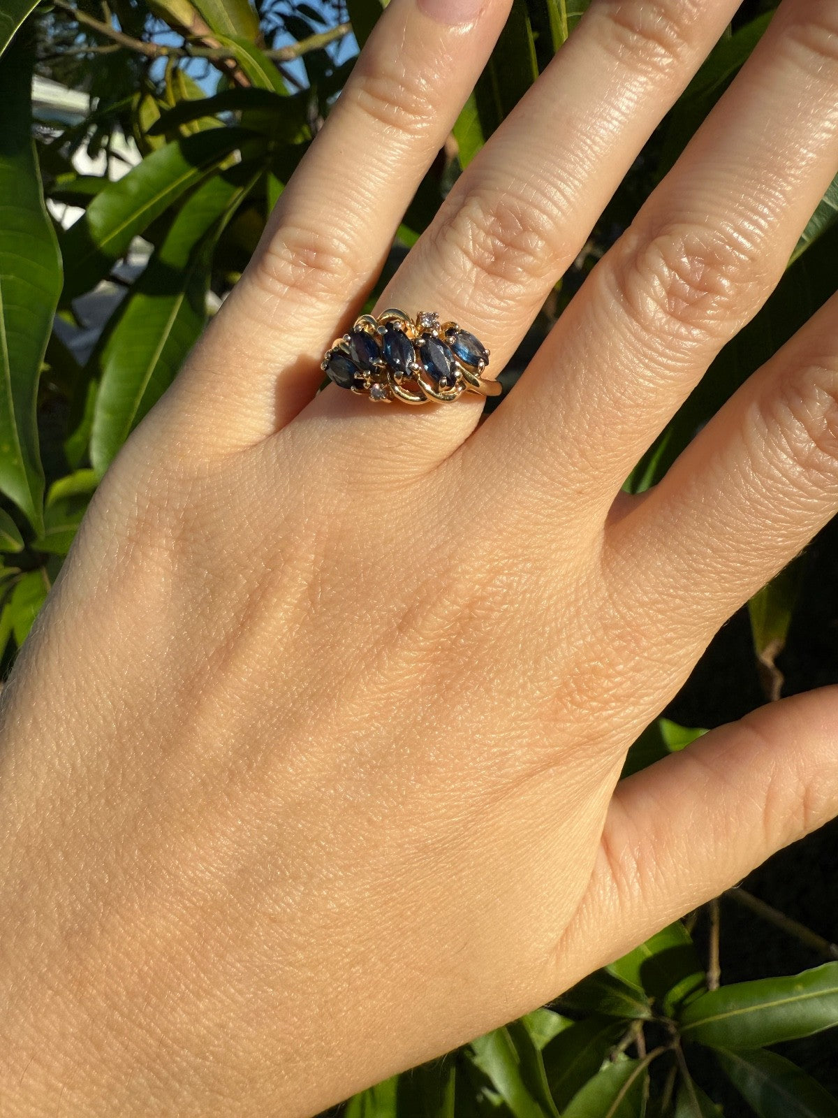 Hand wearing vintage 1960s 14K gold sapphire and diamond swirl bypass ring with marquise blue sapphires, outdoor leafy background.