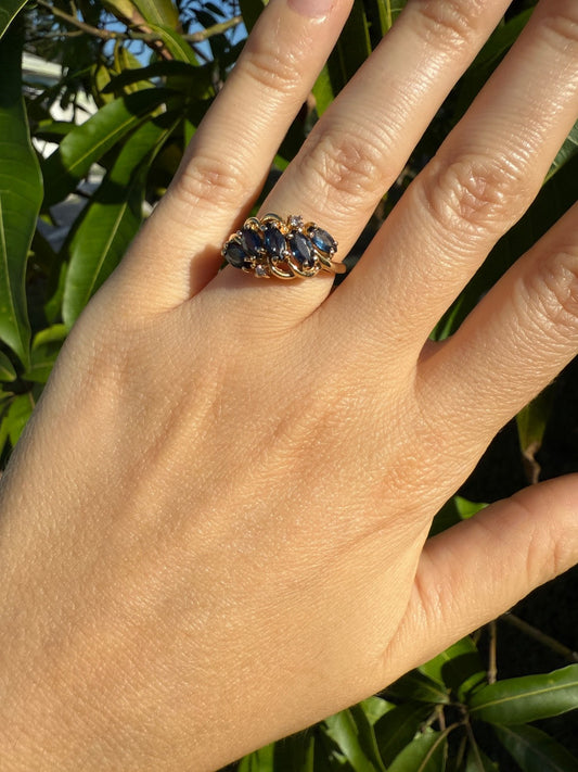 Hand wearing vintage 1960s 14K gold sapphire and diamond swirl bypass ring with marquise blue sapphires, outdoor leafy background.