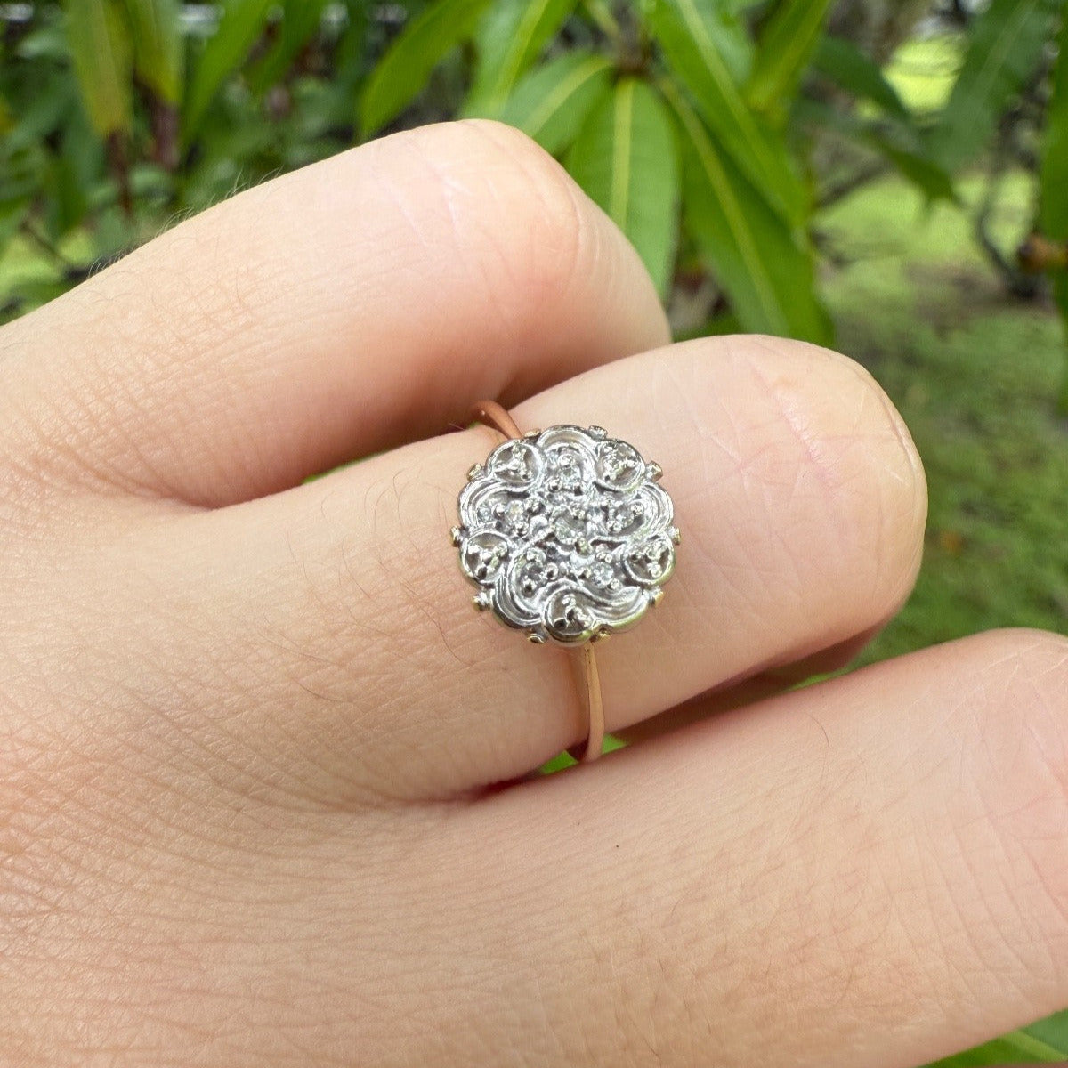 1950s vintage 10K gold diamond illusion cluster ring with ornate flower motif, worn on hand outdoors with green natural background.