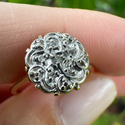 Close-up of 1950s vintage 10K gold diamond illusion cluster ring with ornate flower motif, held between fingers.