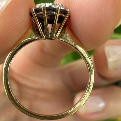 Side profile of 1950s vintage 10K gold diamond illusion cluster ring with slim yellow gold band and ornate basket setting, held between fingers against blurred green background.