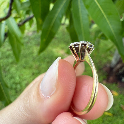 Side profile of 1950s vintage 10K gold diamond illusion cluster ring with ornate basket setting, held between fingers against leafy green background.