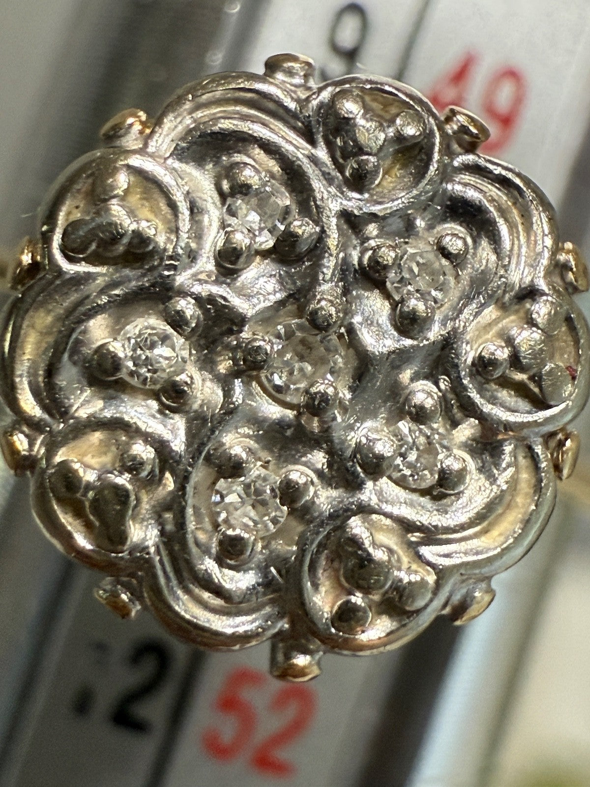Close-up of 1950s vintage 10K gold diamond illusion cluster ring with six diamonds in ornate white gold swirl design, shown on a ring mandrel with size markings in background.