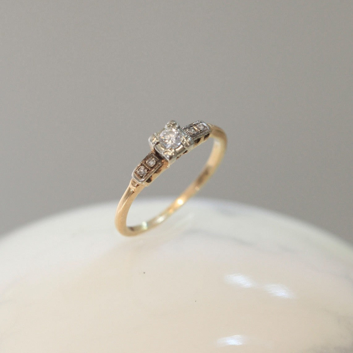 1940s vintage 14K two-tone diamond engagement ring with illusion head and stepped shoulders, displayed on white marble surface.